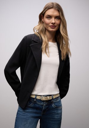 Blonde woman wearing a black jacket, white shirt, beige belt, and blue jeans, standing with hands behind her back against a plain background.