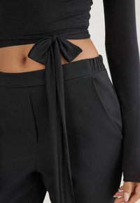 Černý top s dlouhým rukávem a detailem uvázané mašle na zádech. V kombinaci s odpovídajícími černými kalhotami s vysokým pasem, které mají hladkou texturu a sklady.