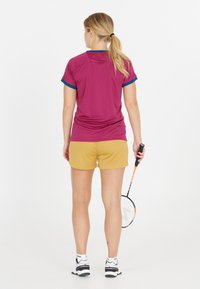 Idrottskläder med en lila T-shirt med kort ärm och blå detaljer samt gula shorts. Personen håller en badmintonracket och har på sig sportskor.