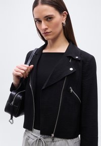 Γυναίκα με καστανά ίσια μαλλιά φοράει μαύρο μπουφάν με φερμουάρ, μαύρο τοπ, γκρι παντελόνι με κορδόνι και κρατάει μαύρη τσάντα πάνω στον ώμο.