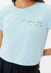 Camiseta de algodón corta, de manga corta y color azul claro, con texto manuscrito en negro que dice: "donde florecen las flores, también florece la esperanza."