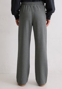 Persoon in losse grijze sweatpants met elastische tailleband en zwart langemouwtop, staat blootsvoets op lichte vloer tegen een witte muur.