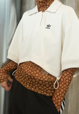 Persoon draagt een cropped witte Adidas polo over een bruine longsleeve top met kleine gele bloemen en zwarte broek met witte strepen.