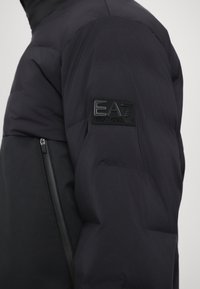 Black puffer jacket featuring a smooth texture and a zippered pocket. Left sleeve displays a black rubber EA7 logo patch.