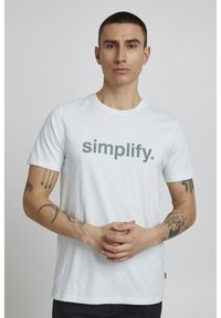 Lyseblå bomulds t-shirt med korte ærmer, der pryder ordet "simplify" i stor grå skrift hen over brystet. Enkelt design, blød tekstur.