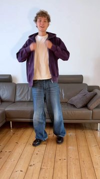 Fialová mikina na zips cez biele tričko, v kombinácii so svetlomodrými širokými džínsami a čiernymi topánkami. Stojí na drevenej podlahe pri šedej pohovke.