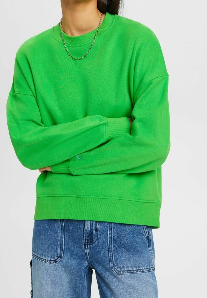 Personne portant un sweat-shirt vert vif à manches longues, bras croisés sur la poitrine, et un jean bleu sur fond blanc.
