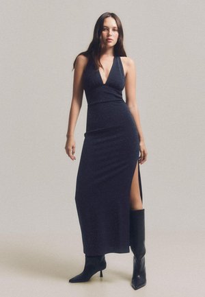 Rochie lungă bleumarin, cu un croi ajustat, decolteu adânc și fenta laterală, realizată dintr-un material neted cu un ușor strălucire. Botine negre cu toc.