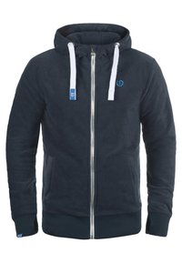 Sudadera con cremallera de forro polar en azul marino oscuro con cordones blancos. Cuenta con dos bolsillos frontales y puños acanalados. Pequeño logo en el pecho.