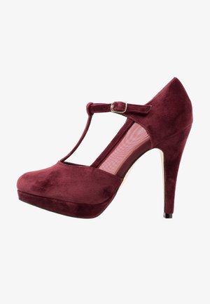 Scarpa con tacco alto in camoscio bordeaux con cinturino a T, punta chiusa, plateau e punta affusolata. Presenta un dettaglio con fibbia argentata.