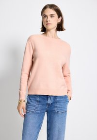Femme aux cheveux bruns courts portant un pull rose clair, un jean en denim bleu avec de grandes poches avant et des bracelets en or, se tenant devant un fond blanc.