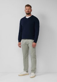Marineblauwe V-hals trui over een wit T-shirt, gecombineerd met lichtgroene broek en witte sneakers met beige zolen. Man staat tegen een neutrale achtergrond.