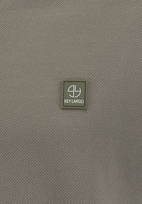Tessuto khaki con una trama a righe diagonali, caratterizzato da una toppa rettangolare verde con il logo e l'icona "KEY LARGO" bianchi.