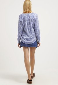 Modrá košile s dlouhým rukávem a bílým květinovým vzorem, zakřiveným spodním lemem a volným střihem, spárovaná s džínsovou sukní a tmavě hnědými botami.