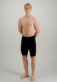 Sorte, tettsittende treningsshorts, lengde til midt på låret. Modellen står barfot med hendene bak ryggen mot en nøytral bakgrunn.