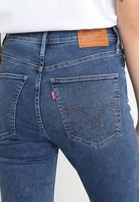 Vaqueros de mezclilla azul oscuro con cintura alta, que presentan bolsillos traseros y una etiqueta de cuero marrón en la parte posterior, con detalles de costura en contraste.