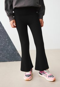Svarta utsvängda byxor i stretchmaterial, kombinerade med färgglada, mönstrade sneakers med rosa, leopardmönster och vita snörningar.
