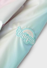 Tissu pastel avec une texture lisse en rose pâle et vert menthe, orné d'un patch en caoutchouc brillant affichant "Unicorn" et un motif arc-en-ciel.