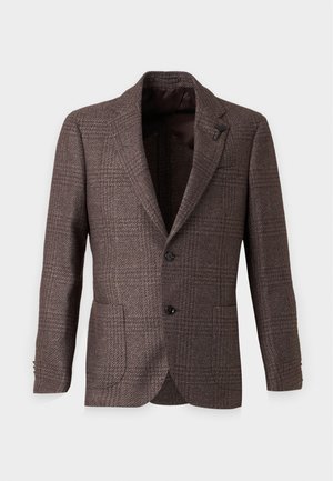 Mann trägt braunen strukturierten Blazer, weißes Knopfhemd, taupefarbene Hose und schwarze Anzugschuhe, steht vor weißem Hintergrund.