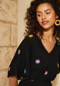 Schwarzes Kleid mit lebhaften, bestickten Sonnenmustern in Gelb, Pink, Grün und Lila. Verfügt über einen V-Ausschnitt und Ärmel mit gewelltem Rand.