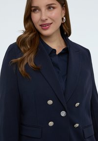 Frau mit langen braunen Haaren, trägt silberne Creolen und einen marineblauen, doppelseitigen Blazer über einem marineblauen Hemd, lächelt leicht und schaut nach rechts.