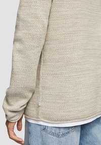 Beige gestrickter Pullover mit einem strukturierten Muster, langen Ärmeln und seitlichen Schlitzen. Getragen über einem weißen Hemd, ergänzt durch hellblaue Jeans.