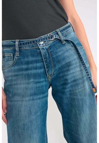 Jeans de denim azul claro con un ajuste relajado, detalles de costura visibles, una bragueta de botón y cremallera, y un cinturón de tela con un lazo.