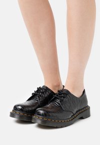 Chaussures à lacets noires brillantes avec motif texturé portées sur pieds nus sur fond blanc.