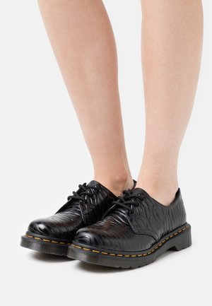 Chaussures à lacets noires brillantes avec motif texturé portées sur pieds nus sur fond blanc.