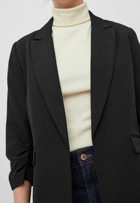 Černý blejzr s klopami, nošený přes světle béžový rolák. Materiál vypadá hladce, s ohrnutými rukávy a detaily kapsy.