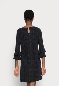 Vestido negro con un patrón de diamantes texturizado, mangas acampanadas y un detalle de ojal en la parte posterior. Tejido suave con un ligero brillo.