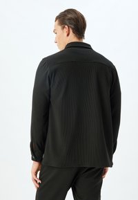 Camicia nera in tessuto testurizzato con colletto, maniche lunghe e dettagli con bottoni sui polsini. Vestibilità rilassata, tessuto morbido, design semplice.