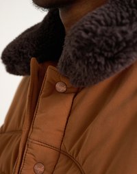 Bruine gewatteerde jas met een donkere bontkraag. Voorzien van drukknopen en een doorgestikte textuur op het lichaam. Close-up van de kraag en schouder.