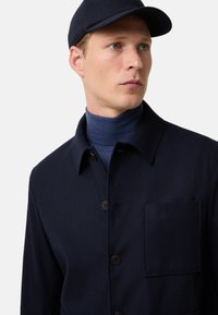 Młody mężczyzna w granatowej czapce, granatowej zapinanej kurtce z kieszenią na piersi i niebieskim golfie, patrzy w prawo na jednolitym tle.