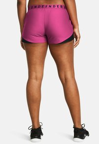 Pantalones cortos deportivos en fucsia con pantalones cortos negros debajo, con una cintura que lleva el texto "UNDER ARMOUR" en negro. Zapatos deportivos negros exhibidos.