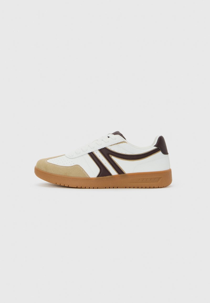 Zapatilla baja blanca con puntera beige, detalles en marrón oscuro y suela de goma marrón, mostrada de perfil sobre un fondo liso.