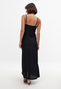 Schwarzes Maxi-Kleid mit dünnen Trägern, strukturiertem Stoff mit dezentem Muster, eng am Oberteil, das zu einem lockeren Saum fließt.