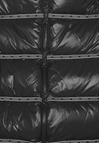 Doudoune noire avec finition brillante, présentant des sections matelassées horizontales et des détails en bandes blanches de la marque le long des coutures.