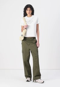 Weißes, kurzes T-Shirt mit gesticktem Text, olivgrüne Cargo-Hosen mit Seitentaschen, beiger Umhängetasche und hellen Sneakers.