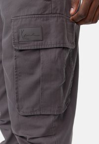 Gray cargo pants with a side pocket, made of durable fabric. Features a rubberized brand patch and visible stitching. Clean, functional design.