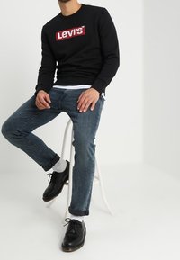 Homme assis sur un tabouret blanc portant un sweat-shirt Levi's noir, un jean bleu, des chaussettes blanches et des chaussures en cuir noir sur un fond blanc uni.