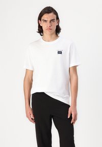 Vit t-shirt med kort ärm och rund halsringning, med en liten svart logotyp på bröstet. I kombination med svarta löst sittande byxor.