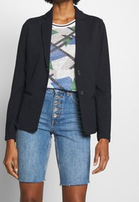 Blazer noir à deux boutons avec poches latérales, associé à un t-shirt gris graphique avec des formes bleues et vertes, et un short en denim bleu clair.