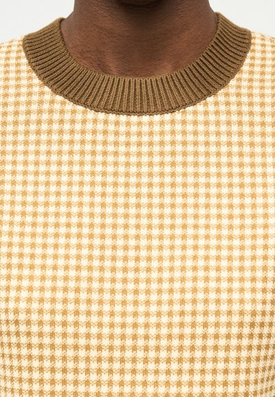 Mørkhudet person som har på seg en beige og lys brun rutete genser med ribbet olivengrønn rund hals.