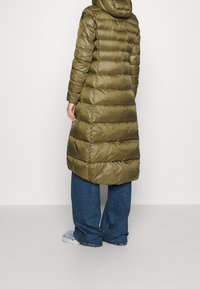 Abrigo tipo puffer color verde oliva con capucha, con diseño acolchado y corte hasta la mitad del muslo. Combinado con jeans azules de pierna ancha y calzado gris.