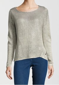 Femme portant un pull en tricot gris clair à manches longues avec un large col rond et un jean bleu foncé.