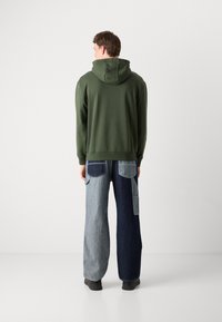 Felpa con cappuccio verde oliva e tasca a marsupio, abbinata a pantaloni a gamba larga in denim bicolore con diverse sfumature e più tasche.