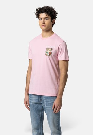 Jonge man staat en draagt een roze T-shirt met een klein afbeelding van een teddybeer en lichtblauwe spijkerbroek tegen een effen witte achtergrond.