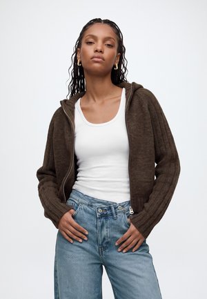 Jeune femme aux longs cheveux tressés portant des boucles d'oreilles en or, un débardeur blanc, un pull marron à fermeture éclair et un jean bleu avec les mains dans les poches.