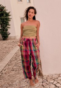 Olívovo zelený naťahovaný top bez ramienok v kombinácii s viacfarebnými károvanými nohavicami v ružových, zelených a modrých odtieňoch. Modelka stojí na kockovanej dlažbe.